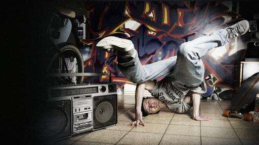 Ein Breakdancer performt auf dem Fliesenboden neben einem Ghettoblaster und vor einer Wand mit Graffitikunst