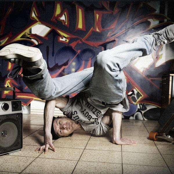 Ein Breakdancer performt auf dem Fliesenboden neben einem Ghettoblaster und vor einer Wand mit Graffitikunst