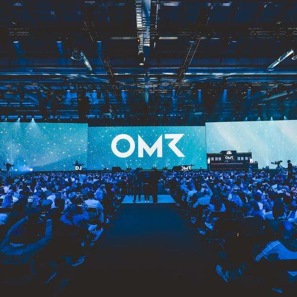 Eine Halle mit einer blau angeleuchteten Bühne und Publikum. Auf der Bühne sieht man die Buchstaben OMR.