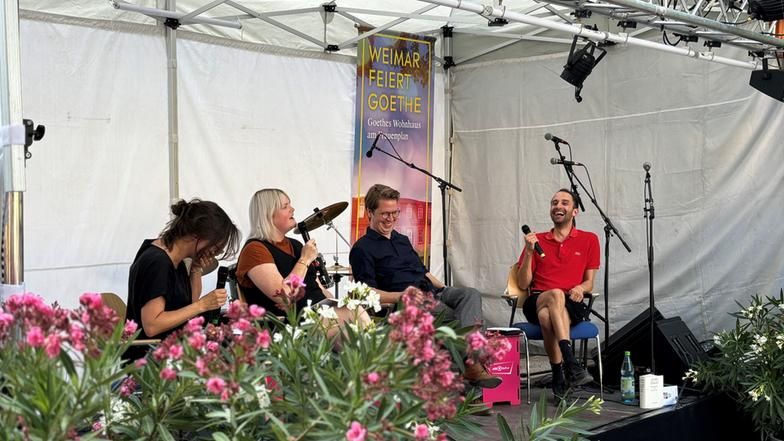 Eine Gruppe von vier Menschen sitzt bei einer Live-Podcast-Aufzeichnung zusammen auf einer Bühne.