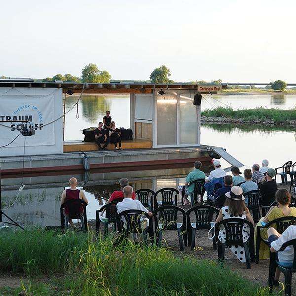 Ein Floß auf dem Wasser steht nah am Ufer, davor einige Reihen an Plastikstühlen. Das Floß dient als Theaterbühne.