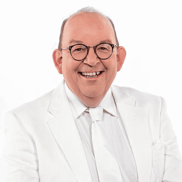 Denis Scheck, ein Mann, mittleren Alters mit Brille und Halbglatze sitzt in weißem Anzug auf einem Tisch.
