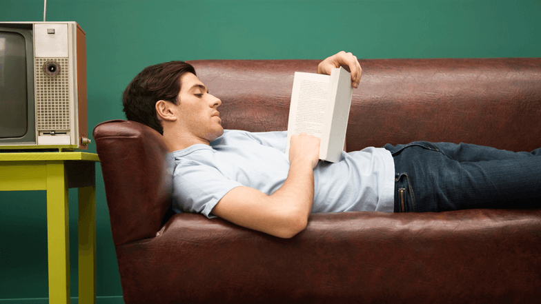 Ein junger Mann liegt lesend auf einem Sofa. Young man relaxing on sofa with book.