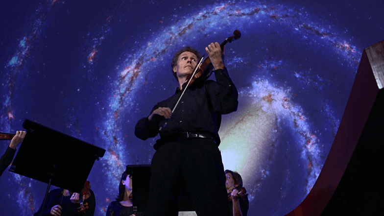 Ein Mann mit Violine steht vor einem Sternenhimmel und spielt auf seinem Instrument.