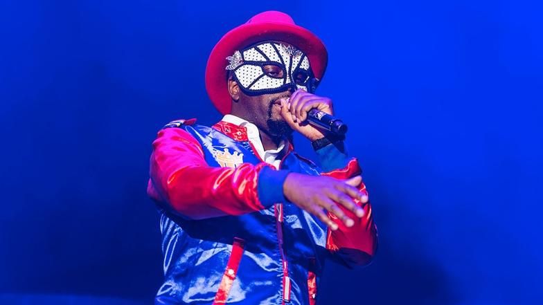 Wyclef Jean auf der Bühne mit Maske, rotem Hut und blauer Jacke.